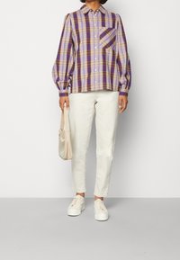 Purple and yellow checkered shirt with puffed sleeves and chest pocket, paired with white jeans and white sneakers, holding a beige bag.