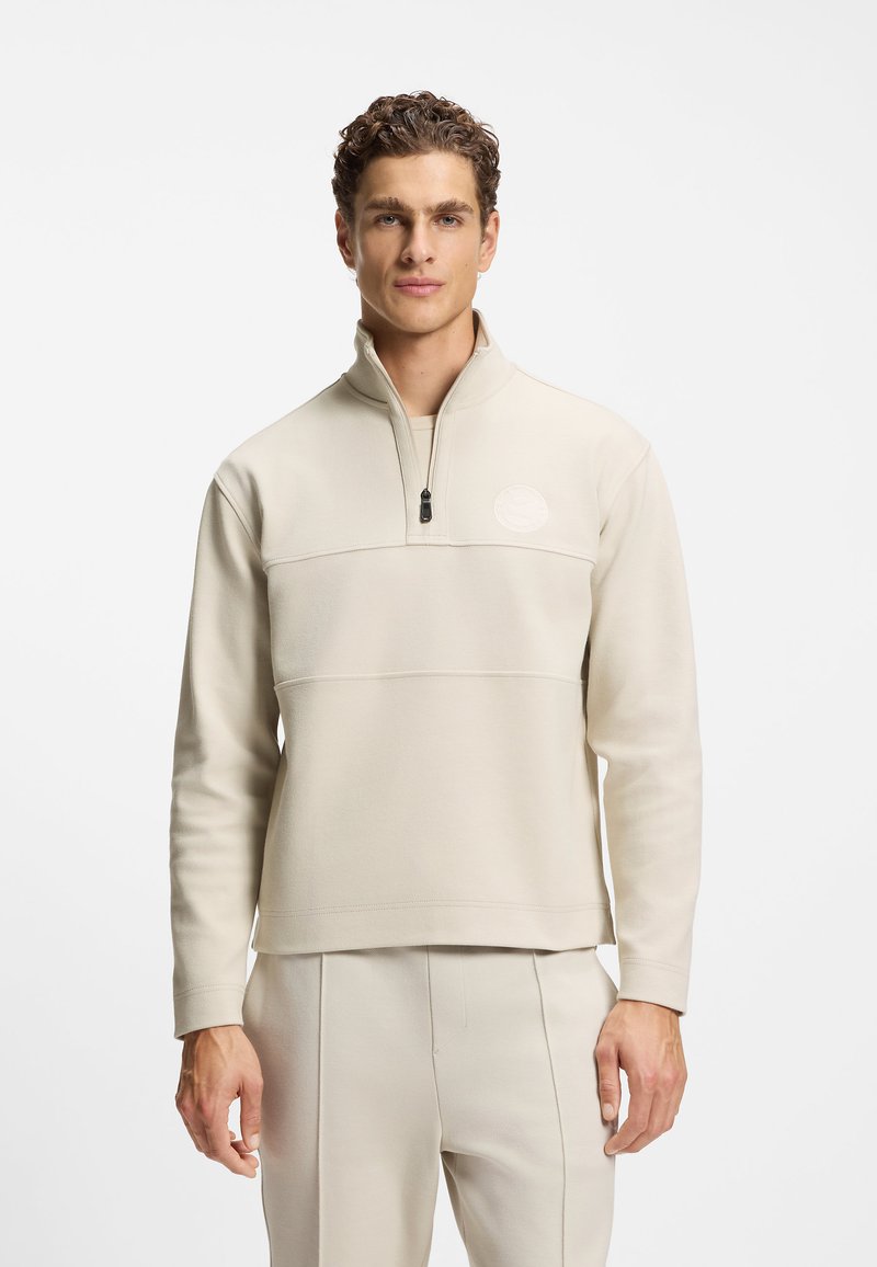 Beżowy pulower z suwakiem do ćwiartki wykonany z miękkiego materiału, z poziomym szwem na wysokości klatki piersiowej oraz okrągłym logo po lewej stronie.