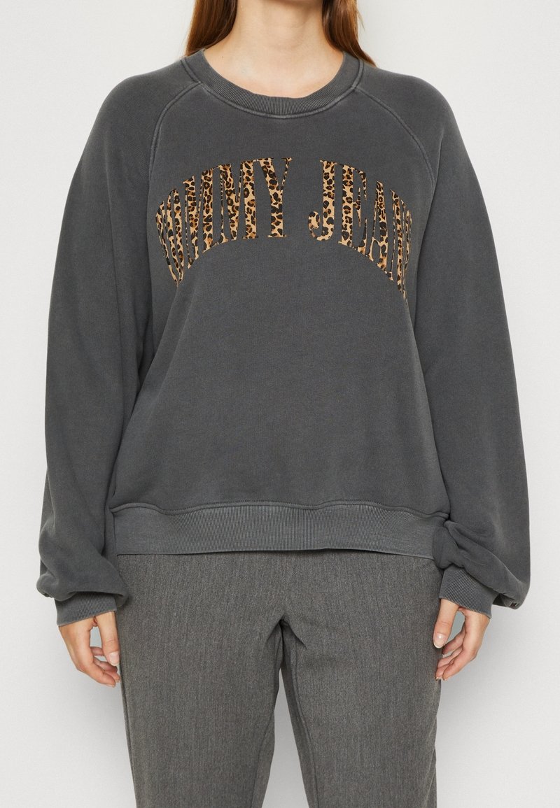 Personne portant un sweat-shirt gris foncé avec "Tommy Jeans" en lettres imprimées léopard et un pantalon gris, debout devant un fond uni.