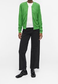 Vihreä pitsinen bomber-takki, jossa on vetoketju edessä, valkoisen paidan yllä, yhdistettynä mustiin leveisiin farkkuihin ja kiiltäviin mustiin nilkkureihin.