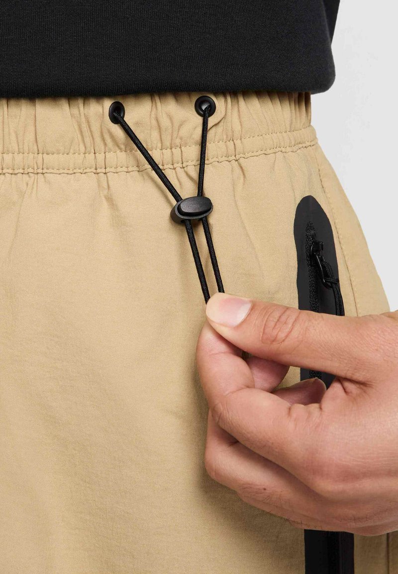 Beige Shorts mit einem elastischen Bund und schwarzem Kordelzug, ausgestattet mit einer Reißverschlusstasche und einem glatten, leichten Stoff. 