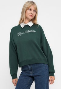 Woman wearing dark green sweatshirt with "Gap Athletic" text, white collared shirt underneath, and blue jeans, posing against plain white background.