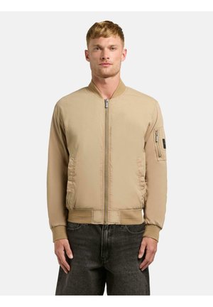 Ung mann som står, iført en beige bomberjakke med glidelås og frontlommer, samt en glidelåslomme på venstre erme, kombinert med mørke jeans.