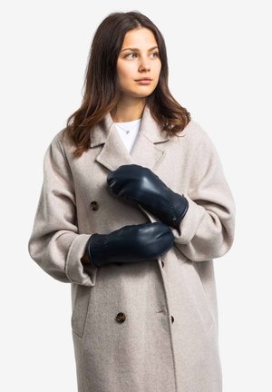 Beige wool coat with a double-breasted design and brown buttons. Dark blue leather mittens. Model stands with hands crossed in front.