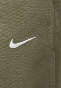 Logotipo da Nike em branco bordado em tecido texturizado verde-oliva, vista aproximada com a costura visível do lado direito.