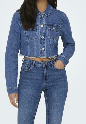 Femme portant une veste en jean bleue courte avec des boutons et un jean bleu taille haute, une main dans la poche et l'autre détendue à ses côtés.