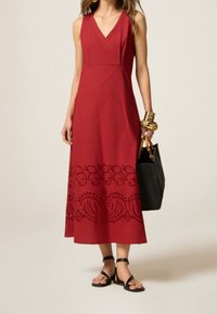 Robe sans manches rouge avec un décolleté en V, ornée de broderies noires délicates au niveau de l'ourlet. Associée à des sandales noires et un sac à main noir texturé.