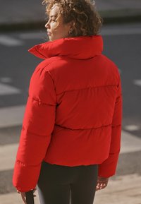 Červená péřová bunda s vysokým golierom, prešívanou textúrou a spadnutými ramennými švami. Model má na sebe tmavé legíny a drží čierny popruh.