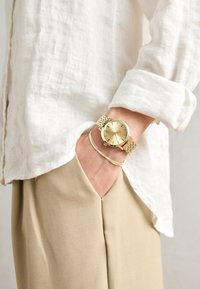Gold wristwatch with a mesh band, round dial, and three black markers. Worn on a wrist alongside a thin gold bracelet. Light-colored fabric visible.