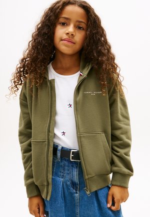 Ung jente med langt krøllete hår iført grønn hettejakke med glidelås, hvit skjorte med stjerner, blå denimskjørt og svart belte, stående foran hvit bakgrunn.