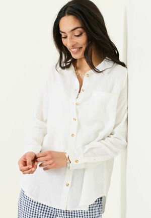 Kvinde med langt mørkt hår iført en hvid skjorte med knapper og blåternet bukser, der smiler og kigger ned.