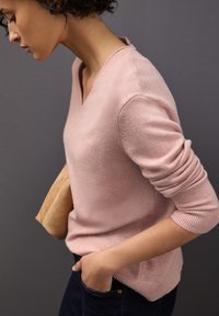 Suéter de punto rosa claro con escote en V, puños y dobladillo acanalados. La modelo lleva jeans oscuros, sosteniendo un clutch beige.