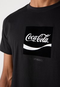 Black T-shirt featuring a prominent white Coca-Cola logo on a square black background, with "ANTONY MORATO" printed below in small text.