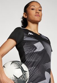 Maillot de sport noir avec des motifs géométriques gris et des manches courtes, tenu avec un ballon de soccer texturé noir et blanc.