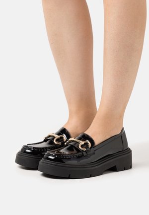 Schwarze Lackleder Slipper mit dicken Sohlen und goldener Pferdebitte, barfuß getragen vor weißem Hintergrund.