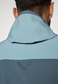 Világoskék kapucnis dzseki sötétkék alsó résszel. Texturált anyaggal rendelkezik, és diszkrét logó részlet található a hátsó részen.