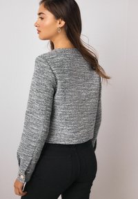 Maglione grigio dalla trama strutturata con design corto e maniche lunghe, caratterizzato da un dettaglio con bottoni sul polsino, abbinato a pantaloni aderenti neri.