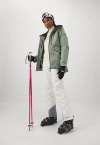 Γυναίκα που φοράει πράσινο μπουφάν, άσπρο σκι παντελόνι, μαύρες μπότες σκι, γάντια και ροζ γυαλιά σκι, κρατώντας δύο κόκκινες μπαστούνες σκι, στέκεται σε άσπρο φόντο.