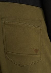 Olijfgroene joggingbroek met een achterzak. Heeft een geribbelde tailleband en een klein bruin logo op de zak, met een gladde stofstructuur.