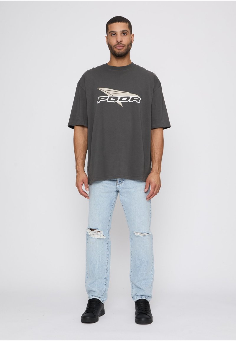 Bărbat purtând un tricou supradimensionat gri închis cu un logo bej, blugi albaștri deschis, rupți, și adidași negri, stând în fața unui fundal alb simplu.