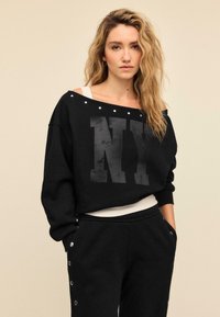 Crna oversized majica s kapuljačom s grafikom "NY", dizajnom koji otkriva ramena, srebrnim detaljima na izrezu oko vrata i rebrastim manšetama. Uparena s crnim trenirkama.