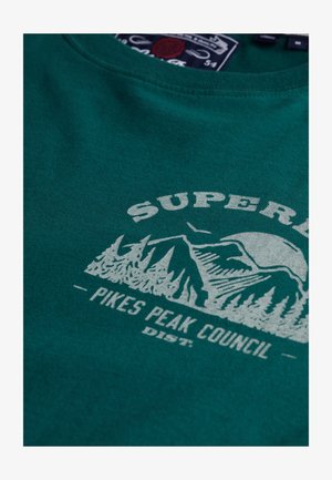 Grøn bomulds t-shirt med et hvidt grafisk design af bjerge og træer, med teksten "SUPER" og "PIKES PEAK COUNCIL" skrevet under.