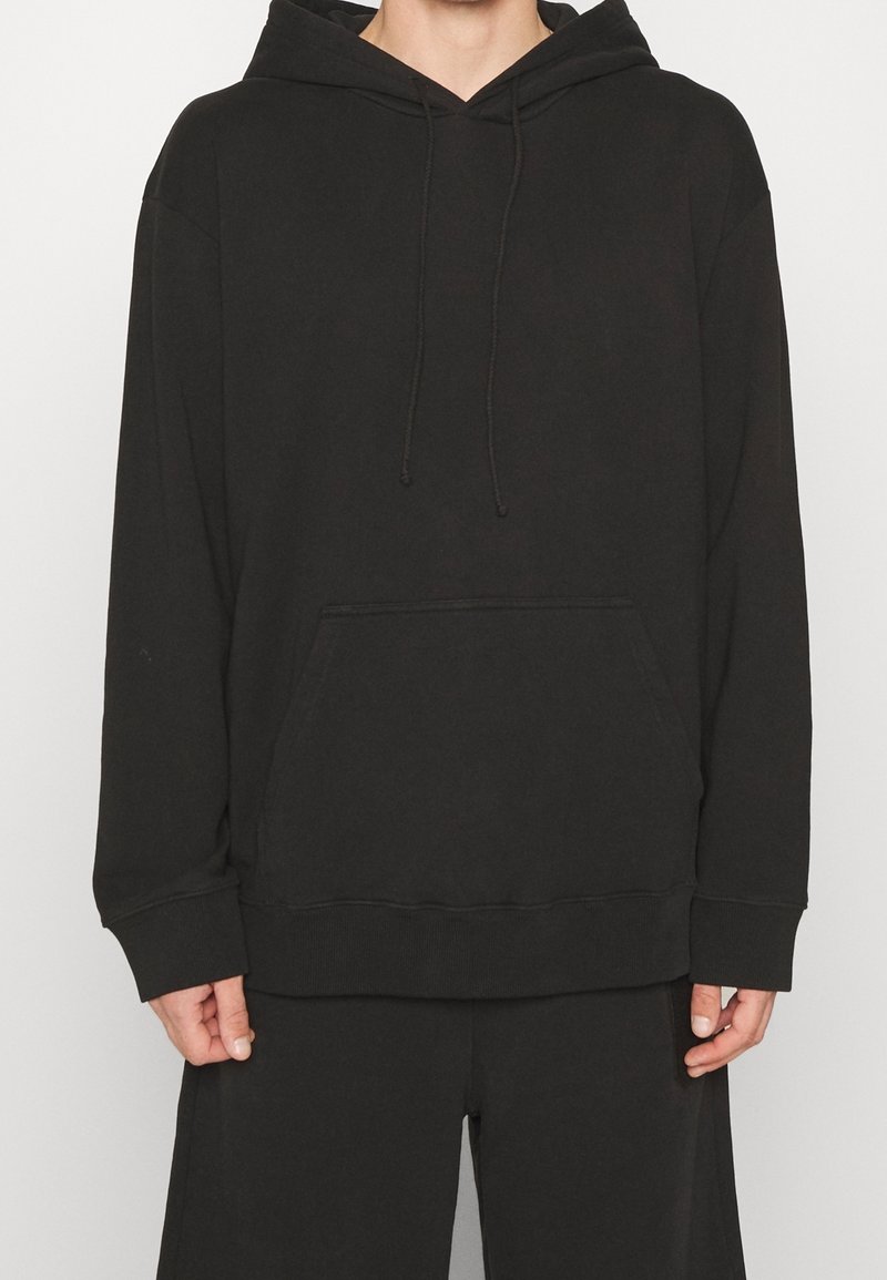 Personne portant un sweat à capuche noir uni avec cordons et poche frontale, associé à un pantalon noir ample, sur fond blanc.