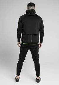 Czarny sportowy hoodie z wysokim kołnierzem, krótszym tyłem i zielonymi akcentami; połączony z czarnymi dopasowanymi spodniami z kieszeniami cargo.