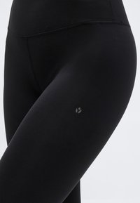 Leggings de sport noirs ajustés à taille haute avec petit logo réfléchissant sur la cuisse, sur fond uni.