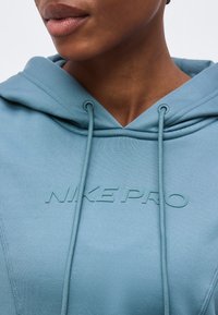 Κοντινό πλάνο ενός ατόμου που φοράει ένα γαλάζιο φούτερ Nike Pro με κορδόνια και ανάγλυφο λογότυπο στο μπροστινό μέρος.