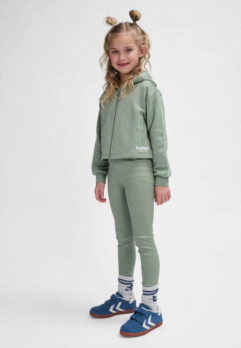Groene bijpassende hoodie- en leggingset in geribbelde textuur; heeft een voorrits en logo; gecombineerd met blauwe schoenen en gestreepte sokken.