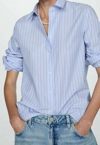 Chemise bleu clair à boutons avec rayures blanches et violettes, dotée d'un col classique et de manches retroussées, assortie à un bas en denim clair.