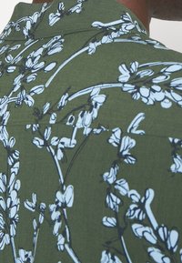 Camicia a maniche corte in tessuto verde oliva con una stampa floreale bianca, colletto rilassato e texture liscia, dettagli di cucitura visibili.