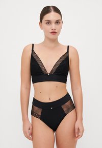 Fekete fehérnemű szett, mély dekoltázsú bralette-tel, hálós részletekkel és magas derekú alsókkal, amelyek hálós akcentusokkal és arany szerelvényekkel díszítettek.