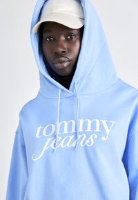 Hoodie bleu clair avec une poche kangourou à l'avant et un logo "tommy jeans" blanc. Accompagné d'une casquette beige avec le nom de la marque brodé.
