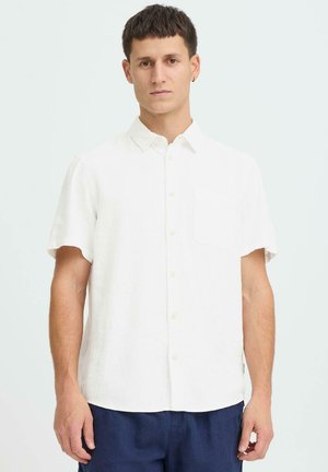 Weißes Leinenhemd mit kurzen Ärmeln, spitzem Kragen, Brusttasche und Knopfverschluss, kombiniert mit dunkelblauer Hose. Glatte Textur.