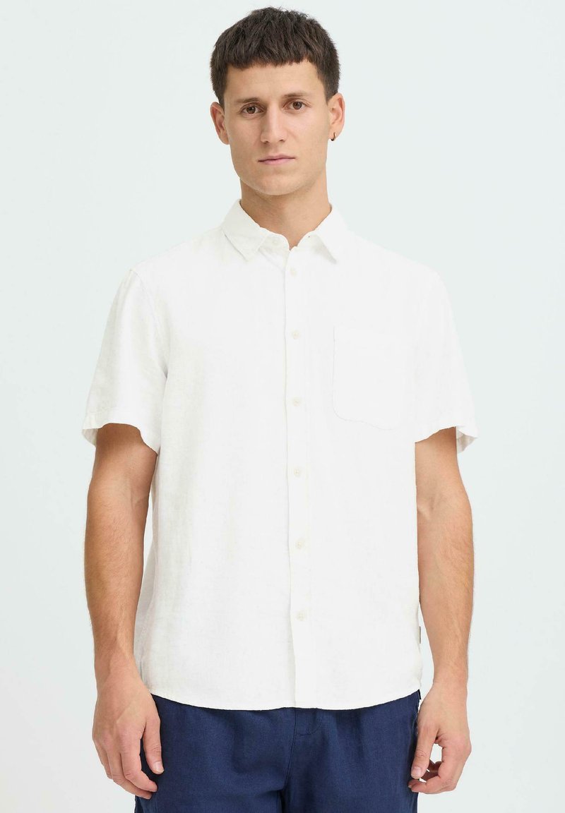 Chemise blanche en lin à manches courtes avec un col pointu, une poche avant et une fermeture à boutons, associée à un pantalon bleu foncé. Texture lisse.