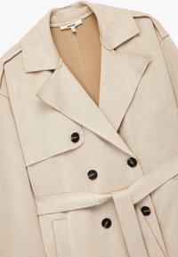 Trench coat de ante sintético beige con cuello con muesca, parte delantera de doble botonadura, botones negros y un cinturón a juego en la cintura.