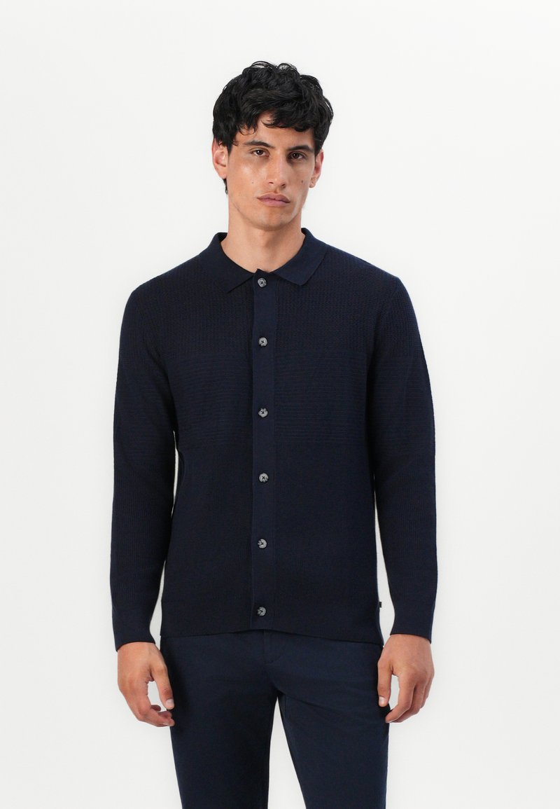 Marineblå langermet cardigan med teksturert strikkemønster, knappestolpe foran og kragehals. Har mørke knapper for lukking.