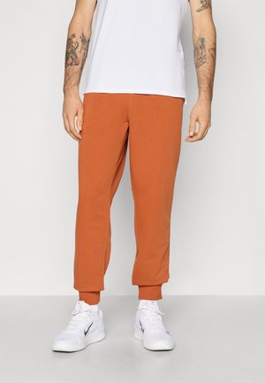 Orange bomuldsjoggers med ribbede manchetter, parret med en hvid T-shirt og hvide sneakers. Stoffet ser blødt og afslappet ud.