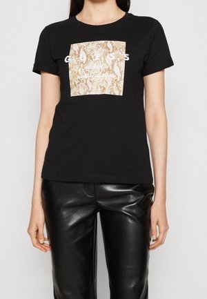 Vrouw draagt een zwart T-shirt met korte mouwen met een beige vierkant met slangenhuidpatroon en witte tekst, gecombineerd met zwarte leren broek.