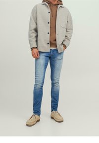 Jasnoszara, teksturowana kurtka na brązowej bluzie z kapturem, zestawiona z wyblakłymi niebieskimi dżinsami i beżowymi butami. Posiada zapięcie na guziki i szczegóły kieszeni.