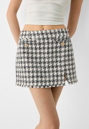 Kvinde iført en høj taljet mini-nederdel i sort-hvidt hundetandsmønster med guldknapper og et lille frontslidt.