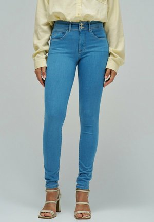 Femme portant un jean skinny taille haute bleu clair, des talons beiges à lanières et une chemise ample boutonnée jaune pâle.