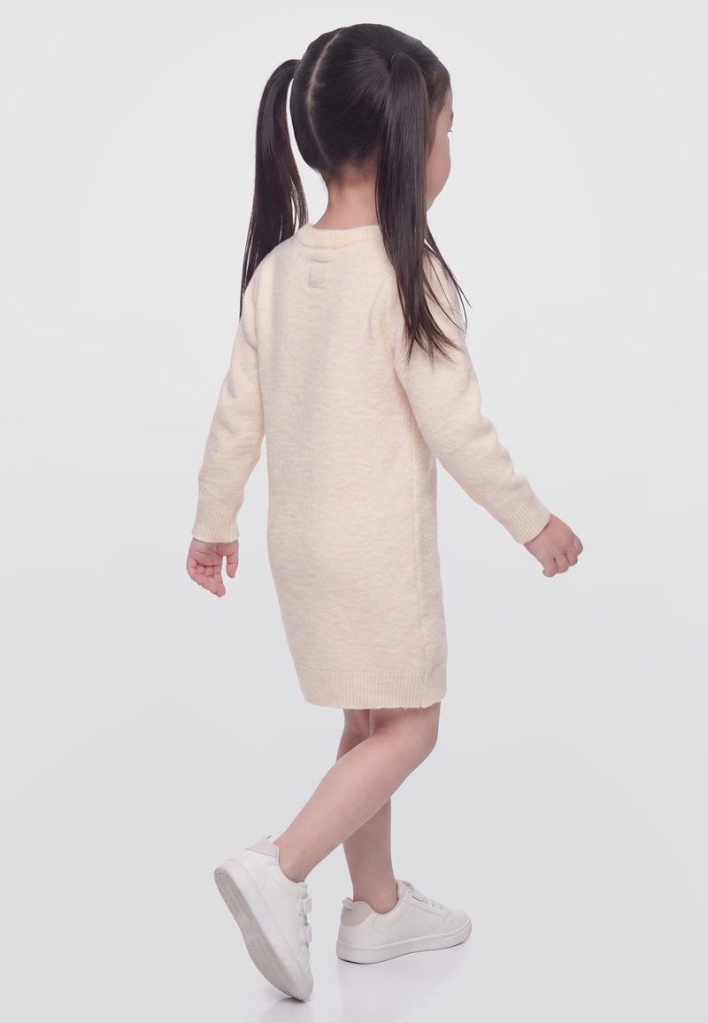 Jeune fille aux longs cheveux foncés en couettes, portant une robe beige à manches longues et des baskets blanches, marchant devant un fond clair uni.