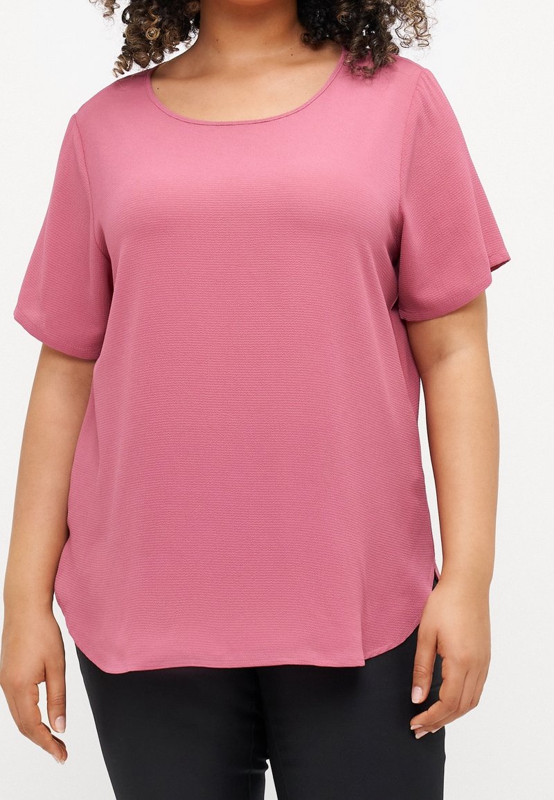 Femme portant un chemisier texturé rose poudré à manches courtes et un pantalon noir, vue de la poitrine aux hanches sur fond blanc.