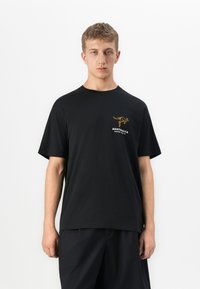 Ung mann iført en svart T-skjorte med en liten oransje kenguru og teksten "Australia Ripcurl Surf Co" på venstre bryst, stående mot en hvit bakgrunn.