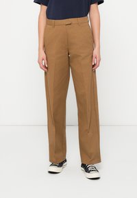 Άτομο με καφέ παντελόνι ίσιας γραμμής, ναυτικό πουκάμισο και μαύρα χαμηλά αθλητικά παπούτσια, που στέκεται απέναντι από έναν απλό λευκό τοίχο.