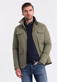 Giacca verde oliva con quattro tasche anteriori, chiusure con zip e bottoni a pressione, colletto alto, indossata sopra un maglione blu navy e una camicia a quadri, abbinata a jeans blu.