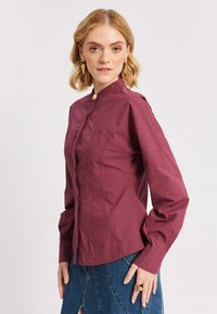 Donna bionda che indossa una blusa attillata a maniche lunghe color bordeaux e una gonna in denim blu, in posa con la mano sui fianchi su uno sfondo chiaro.